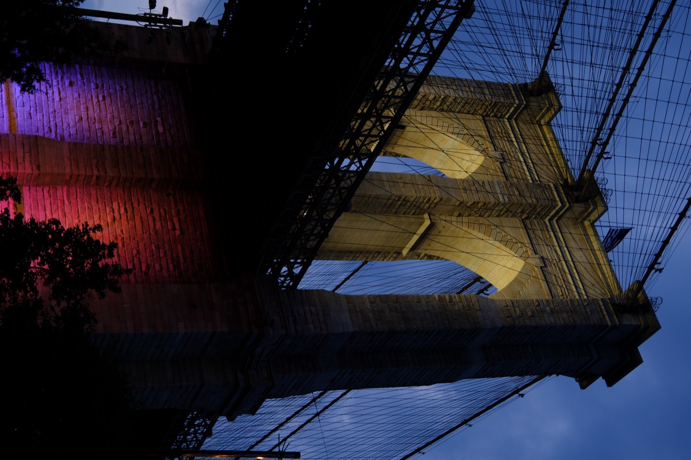 Brooklyn Bridge tower lit up at dusk with colored uplighting on the stonework
